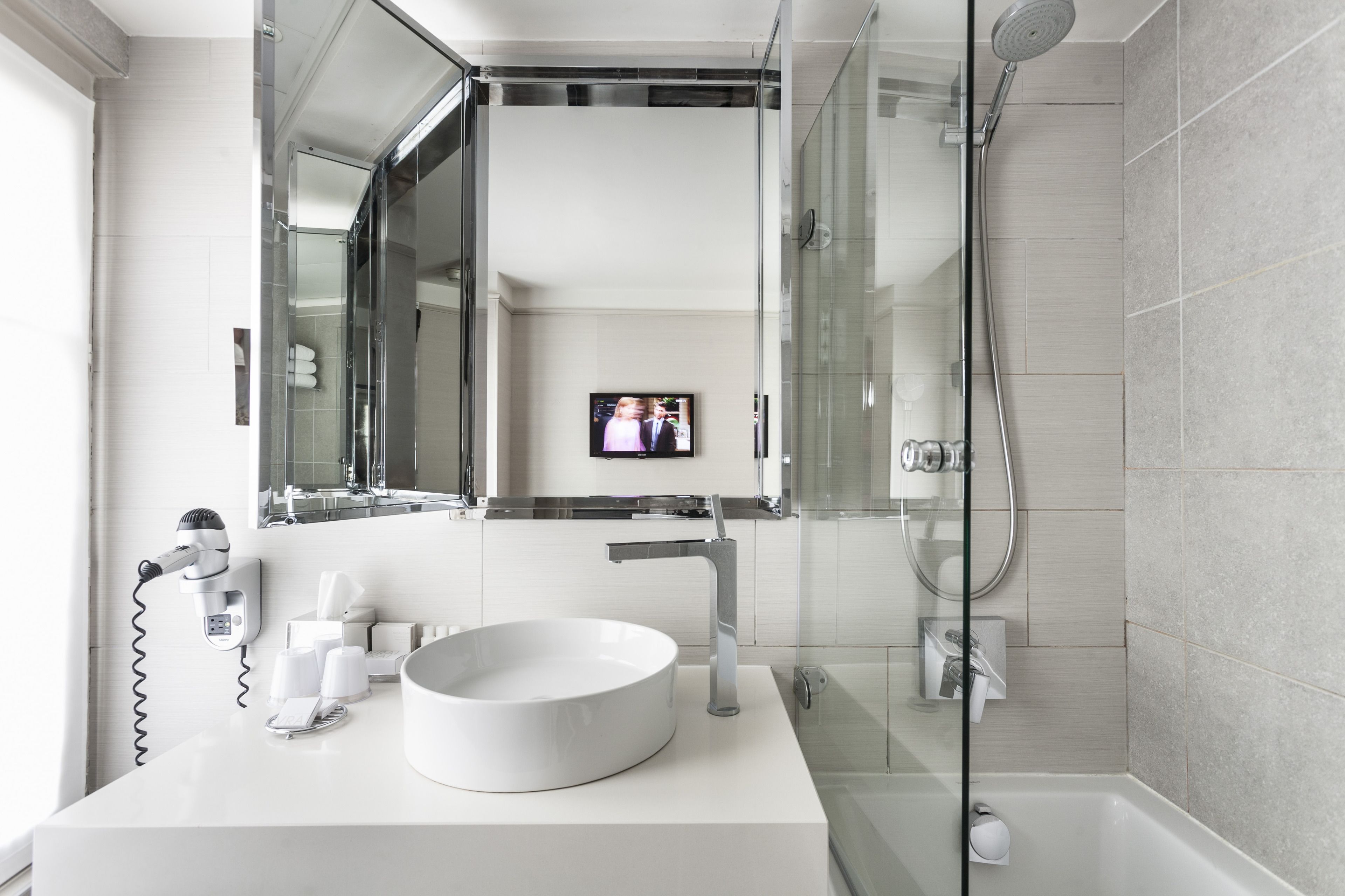 A modern bathroom with a large mirror, a round sink, a handheld showerhead, and a television mounted on the wall.