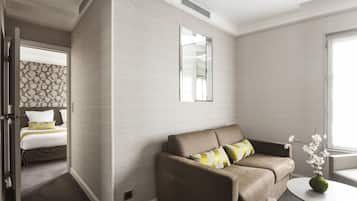 Una habitación de hotel moderna con un sofá beige, una mesa redonda con un florero y una cama con ropa de cama estampada.