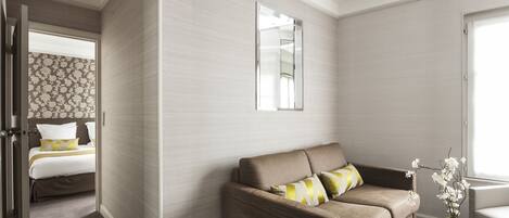 A modern hotel room with a beige sofa, a round table with a vase of flowers, and a bed with patterned bedding.