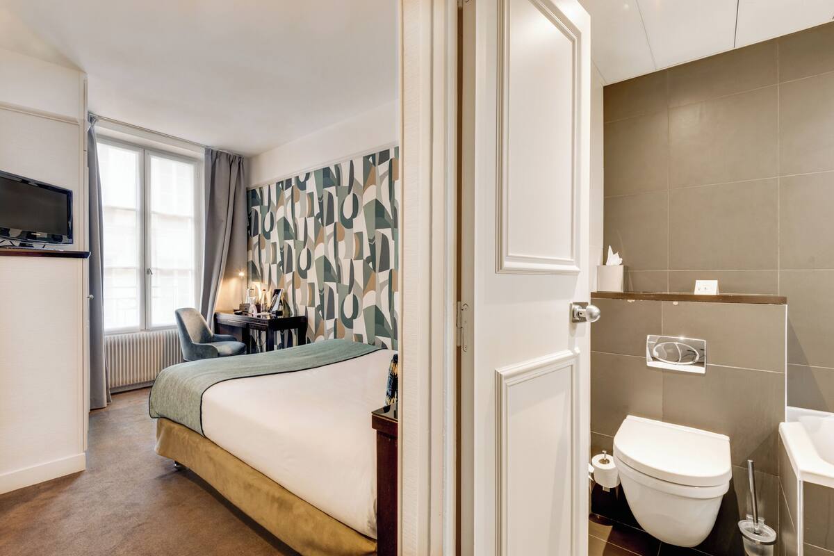 A hotel room with a bed, a desk, a chair, a TV, a bathroom with a toilet and sink.