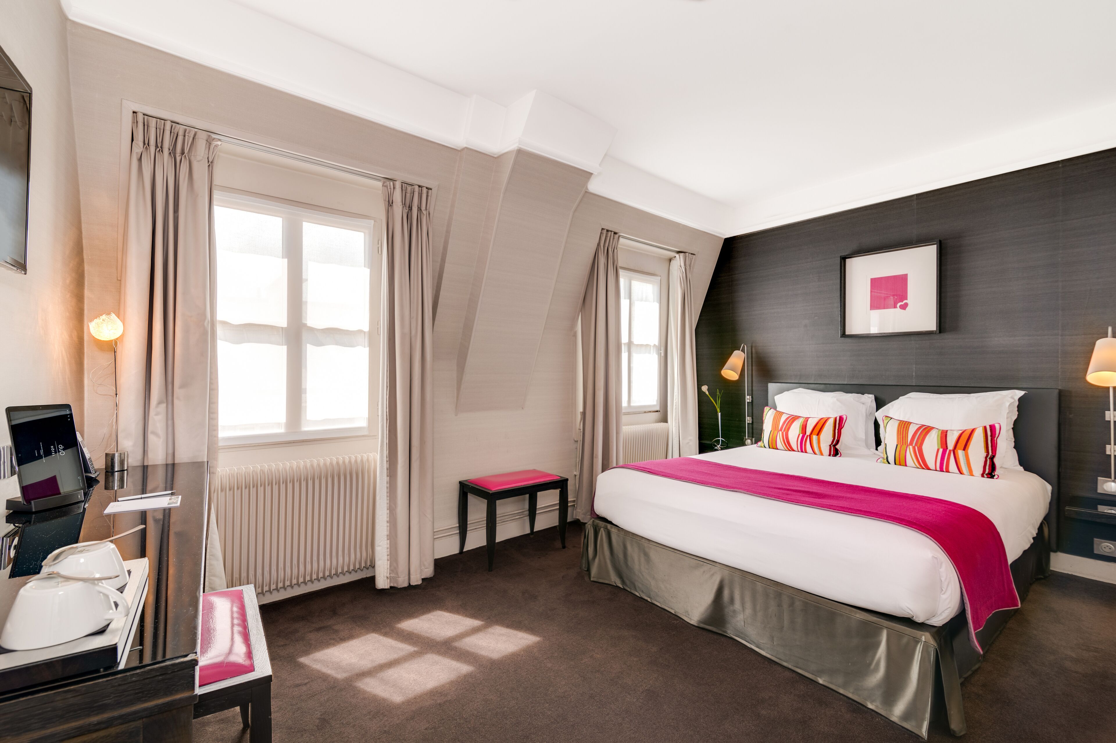 A hotel room with a large bed, a desk with a phone and a laptop, a small table, and a radiator.