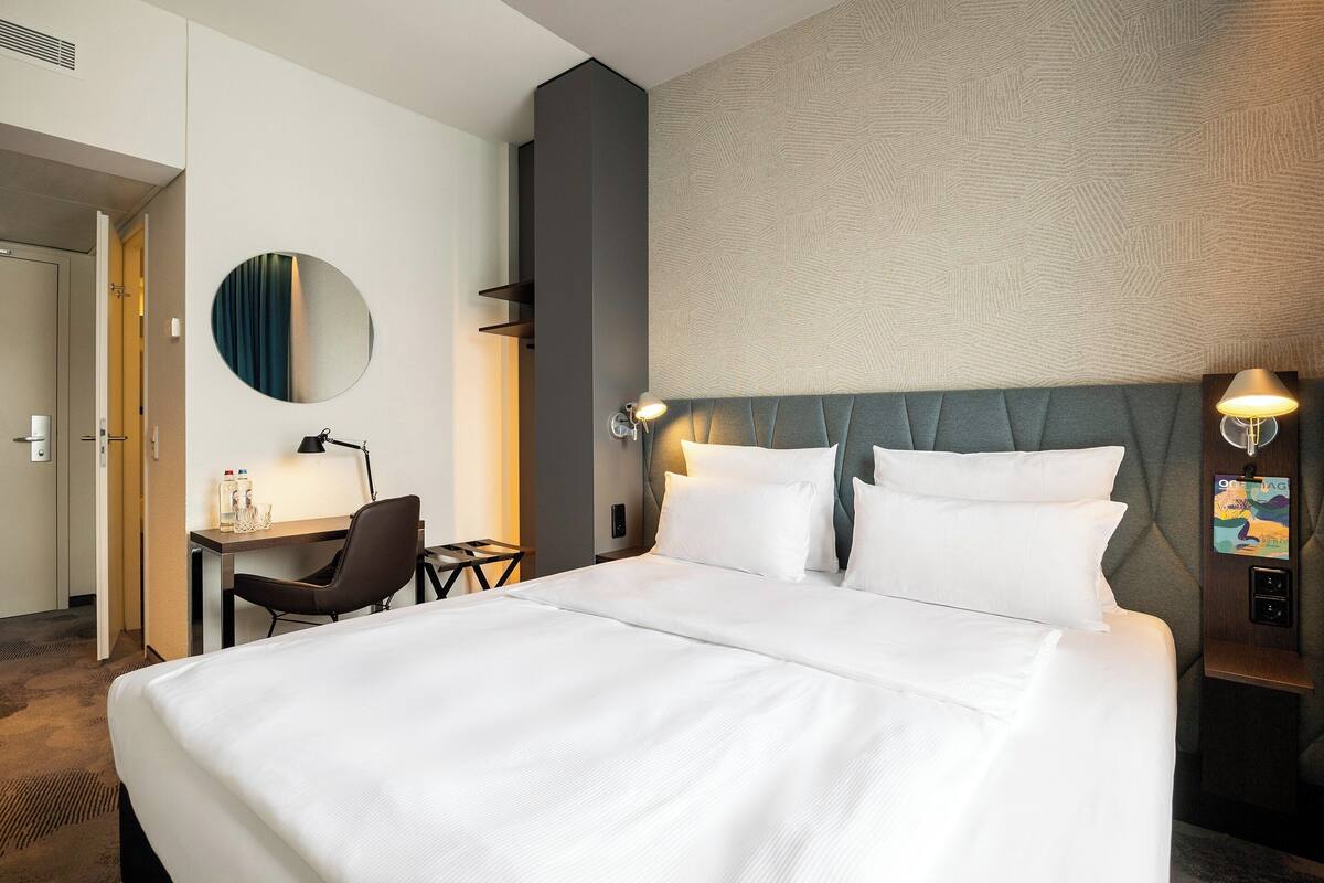 Une chambre d’hôtel avec un grand lit, un bureau avec une chaise, un miroir rond et une tête de lit dotée d’un luminaire.