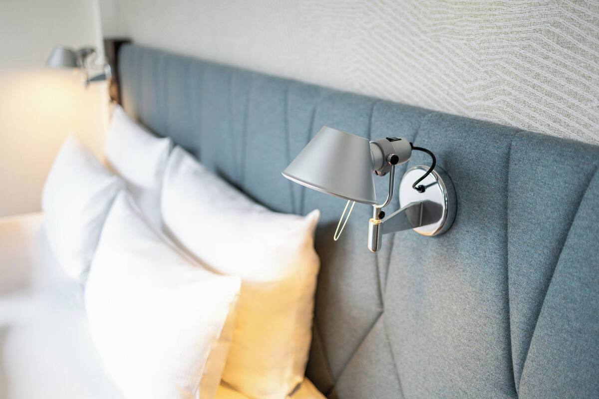 Un lit avec une tête de lit grise, une lampe fixée au mur et des oreillers.