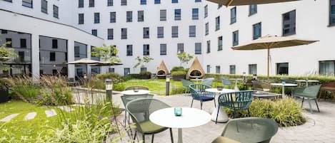 A courtyard with outdoor seating, including tables and chairs, surrounded by greenery and a building with multiple windows in the background.