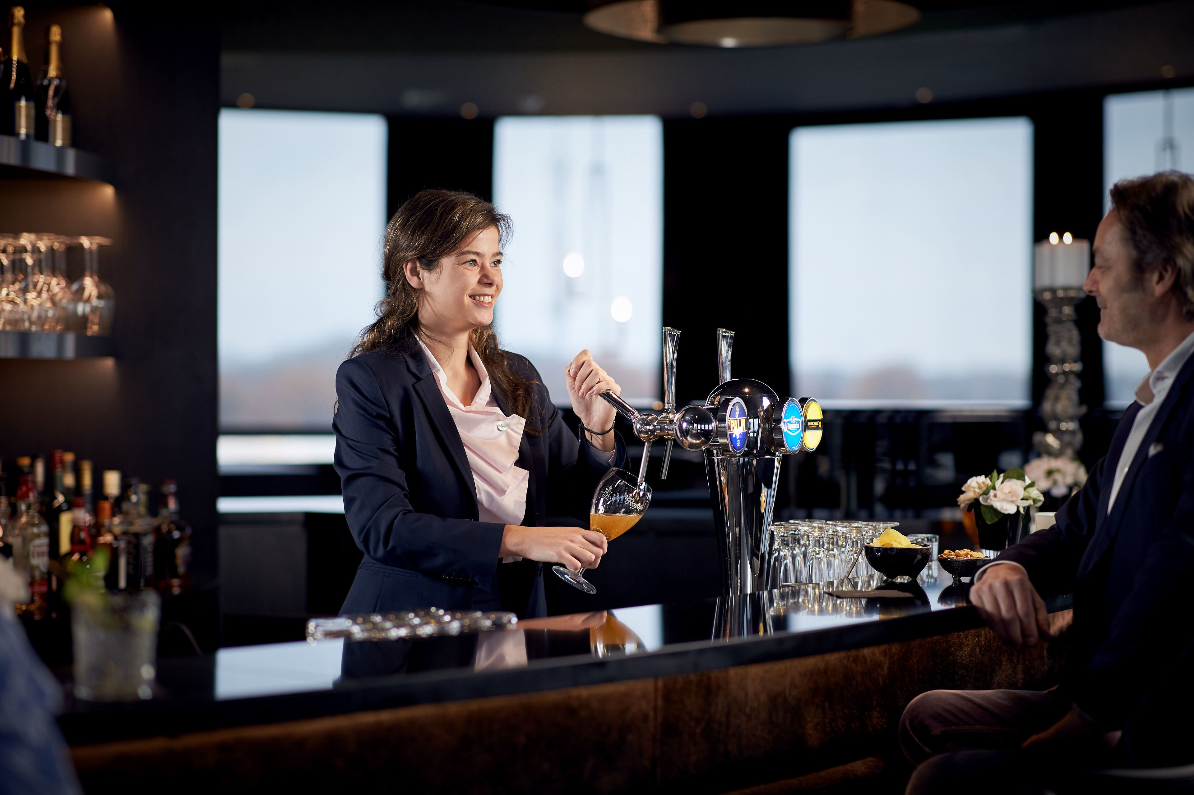 A bar area with a bartender serving a drink, a counter with glasses, and a shelf with bottles.