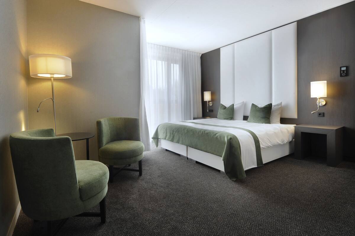 A hotel room with a bed, two chairs, a small table, and wall-mounted lamps.