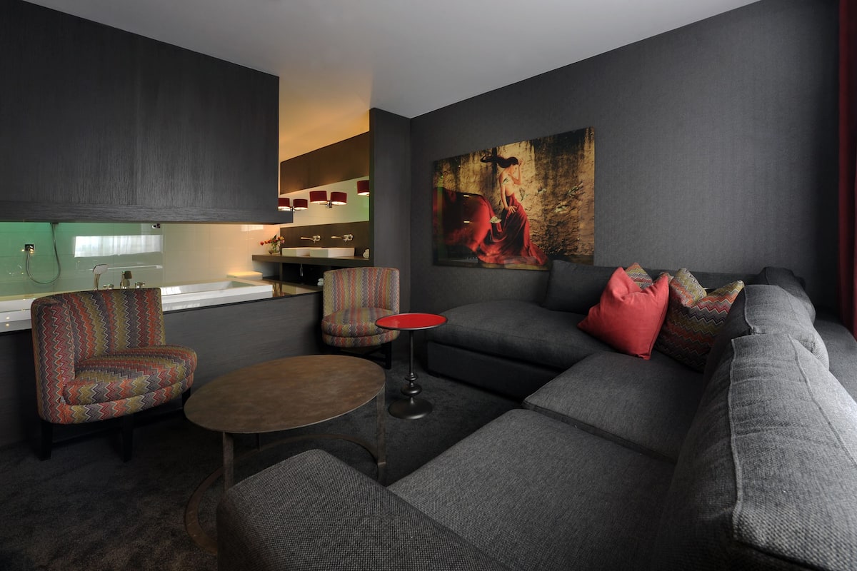A modern living room with a gray sofa, colorful armchair, and a small round table. A painting is on the wall, and a kitchen area is visible in the background.