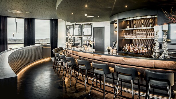 A modern bar with a curved counter, bar stools, and a wine display. - Van der Valk Hotel Uden-Veghel (Uden)
