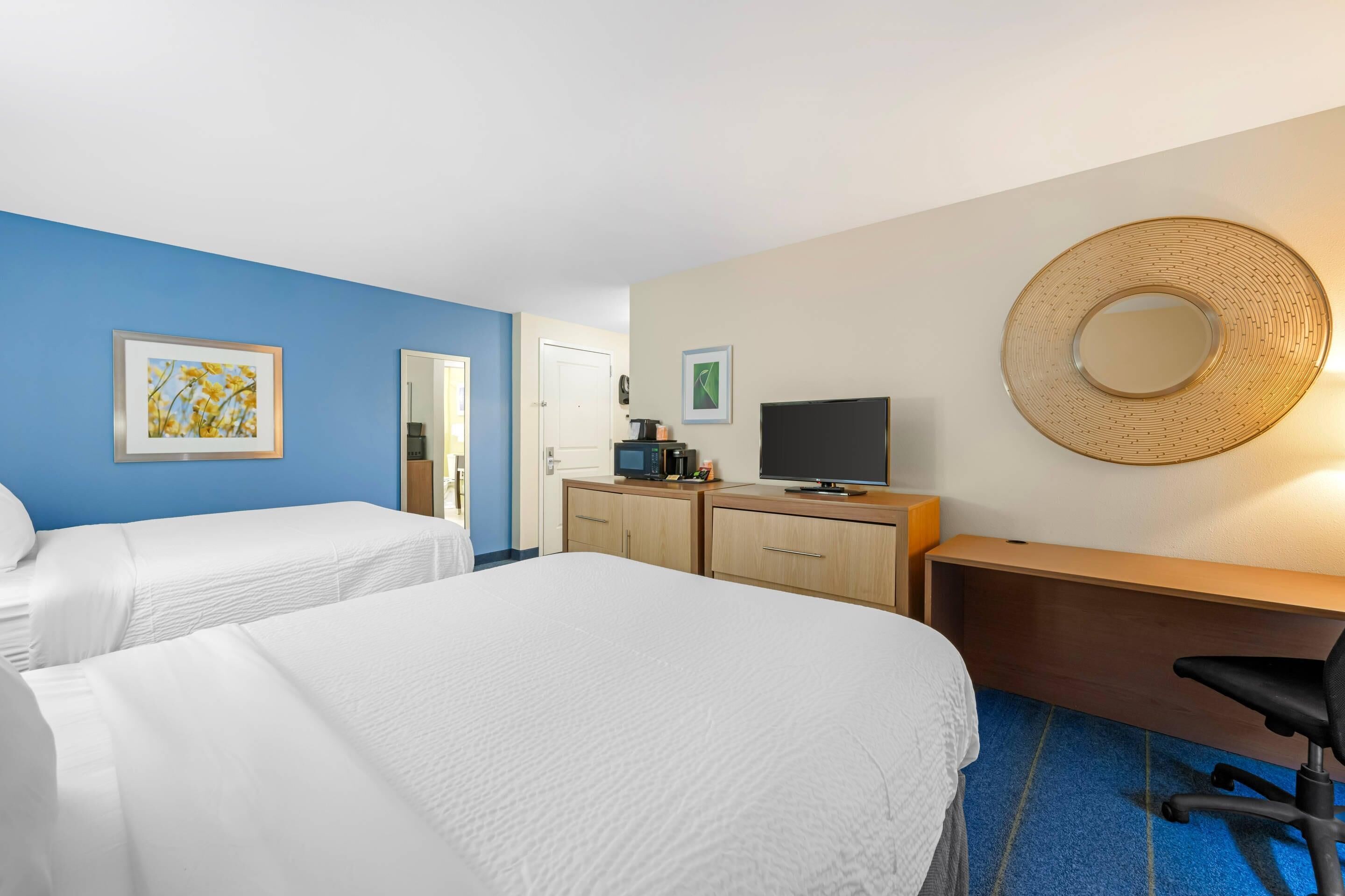 Habitación de hotel con dos camas, un escritorio, una silla, un televisor y un espejo montado en la pared.