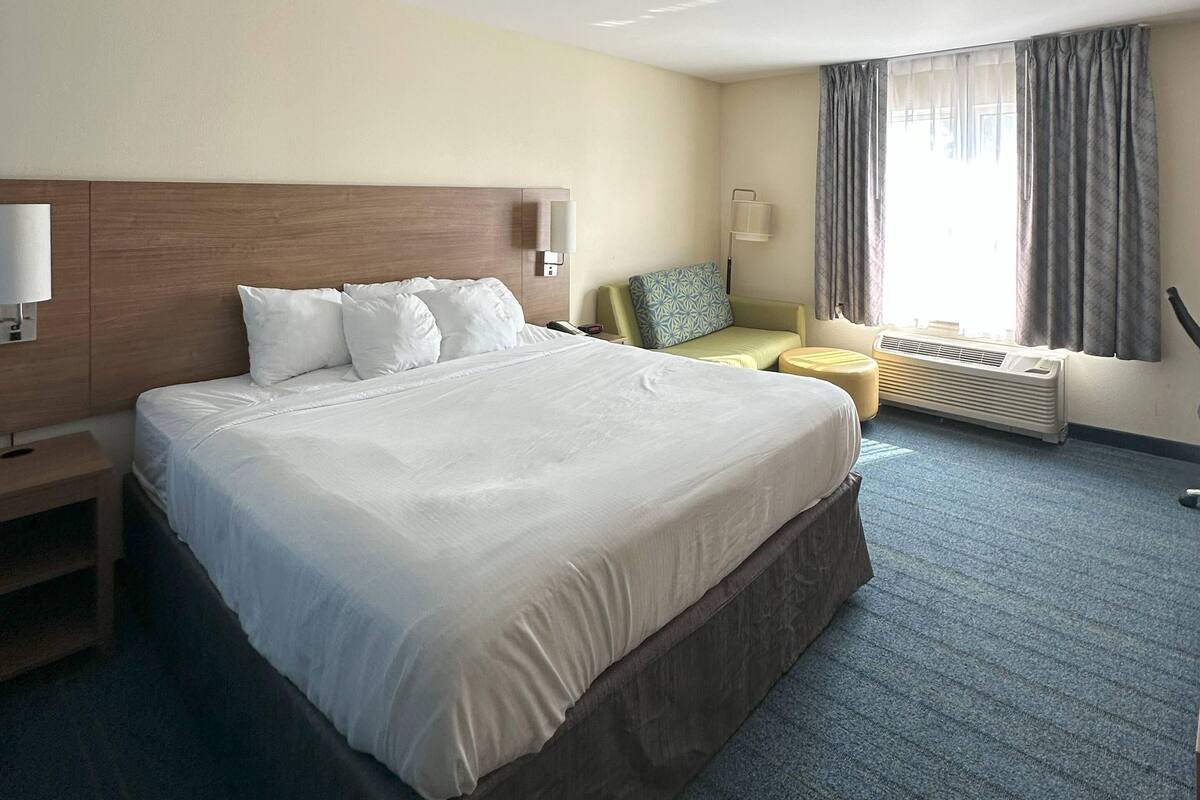 Ein Hotelzimmer mit einem großen Bett, Nachttischen, einem Schreibtisch, einem Sessel, einer Lampe und einem Vorhangfenster.
