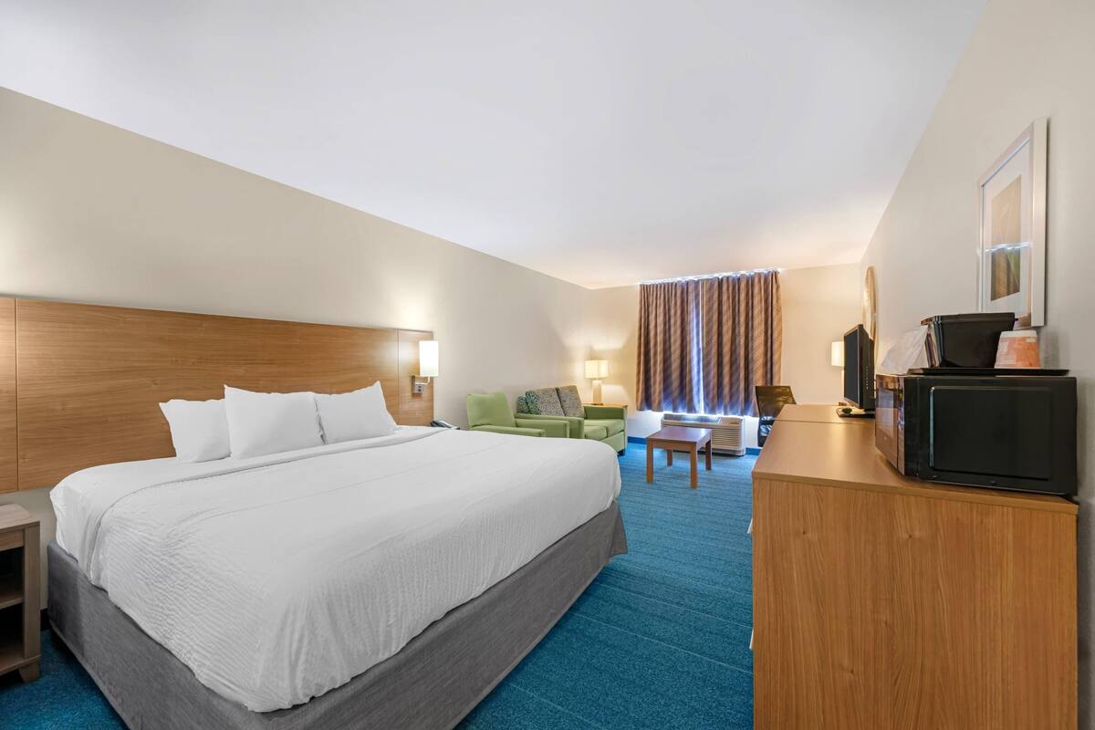 Ein Hotelzimmer mit einem großen Bett, einem Schreibtisch mit Computer, einem Stuhl, einer Lampe und einem Fernseher.