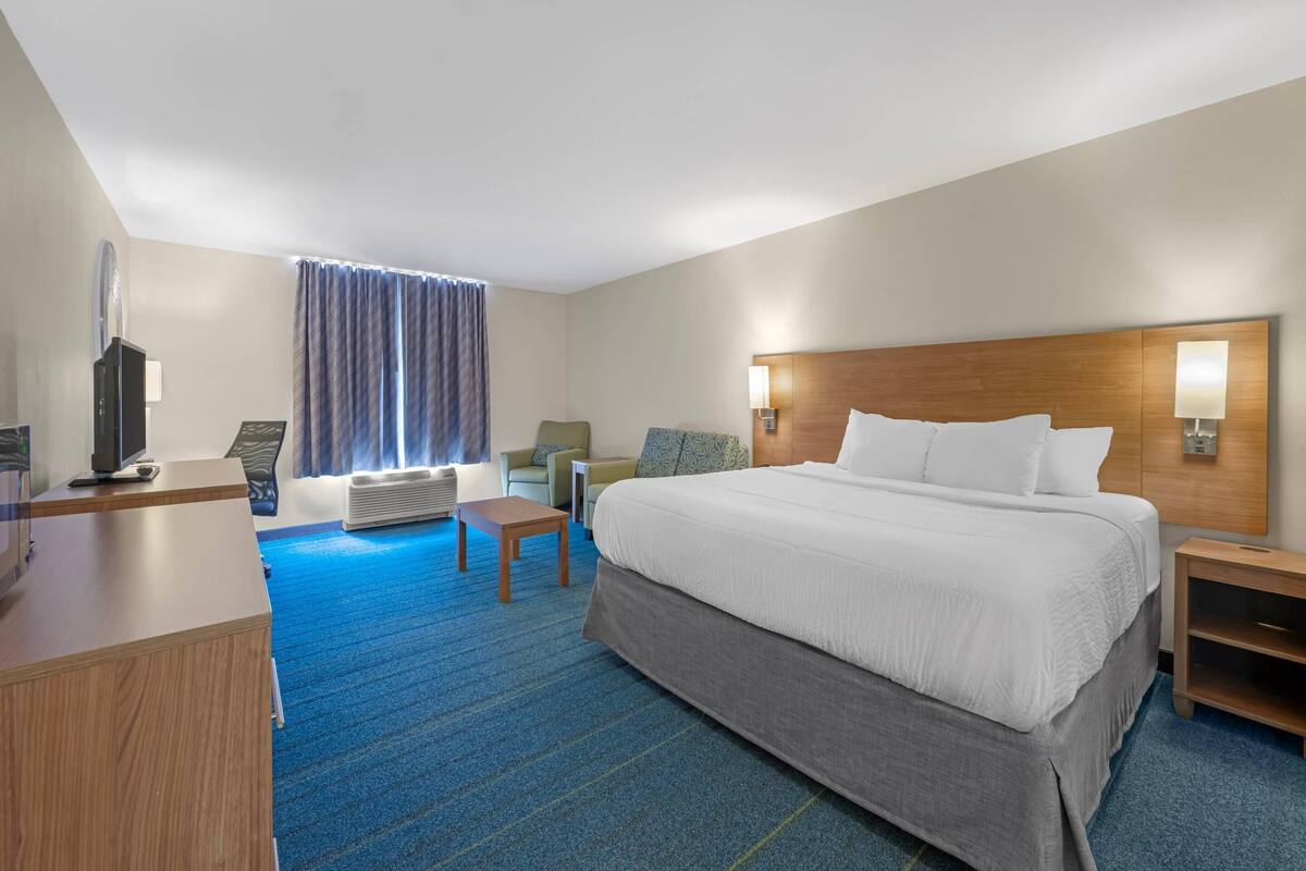 Ein Hotelzimmer mit einem großen Bett, einem Schreibtisch, einem Sessel, einem Fernseher und einem Fenster mit Vorhängen.