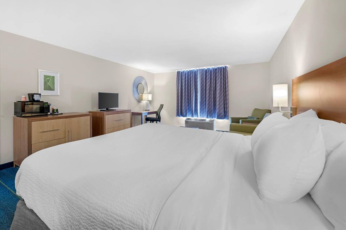 Ein Hotelzimmer mit einem großen Bett, einem Schreibtisch mit Computer, einem Fernseher, einer Lampe und einem Fenster mit Vorhängen.
