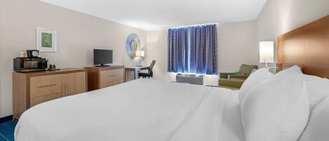 A hotel room with a large bed, a desk with a computer, a TV, a lamp, and a window with curtains.