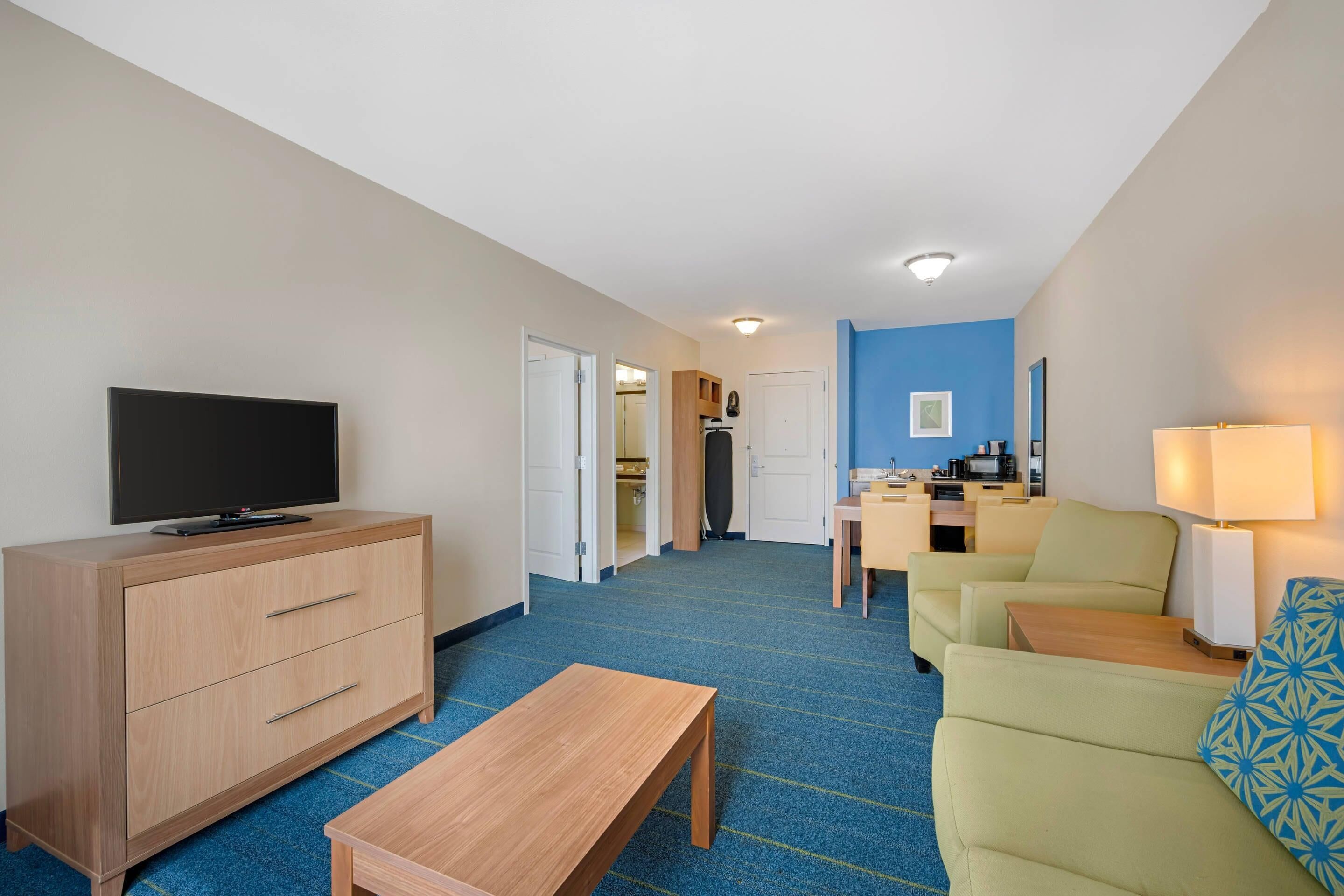 Habitación de hotel con televisión, muebles de madera, alfombra azul, kitchenette y una pared de acento azul.