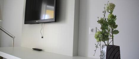 Un televisor de pantalla plana montado en la pared, una mesa blanca con un control remoto y un florero con una planta.