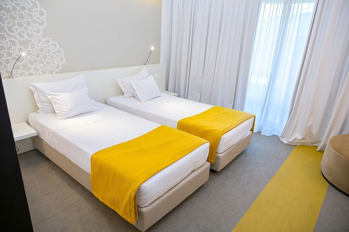 A hotel room with two beds, a bedside table, a lamp, and a window with curtains.