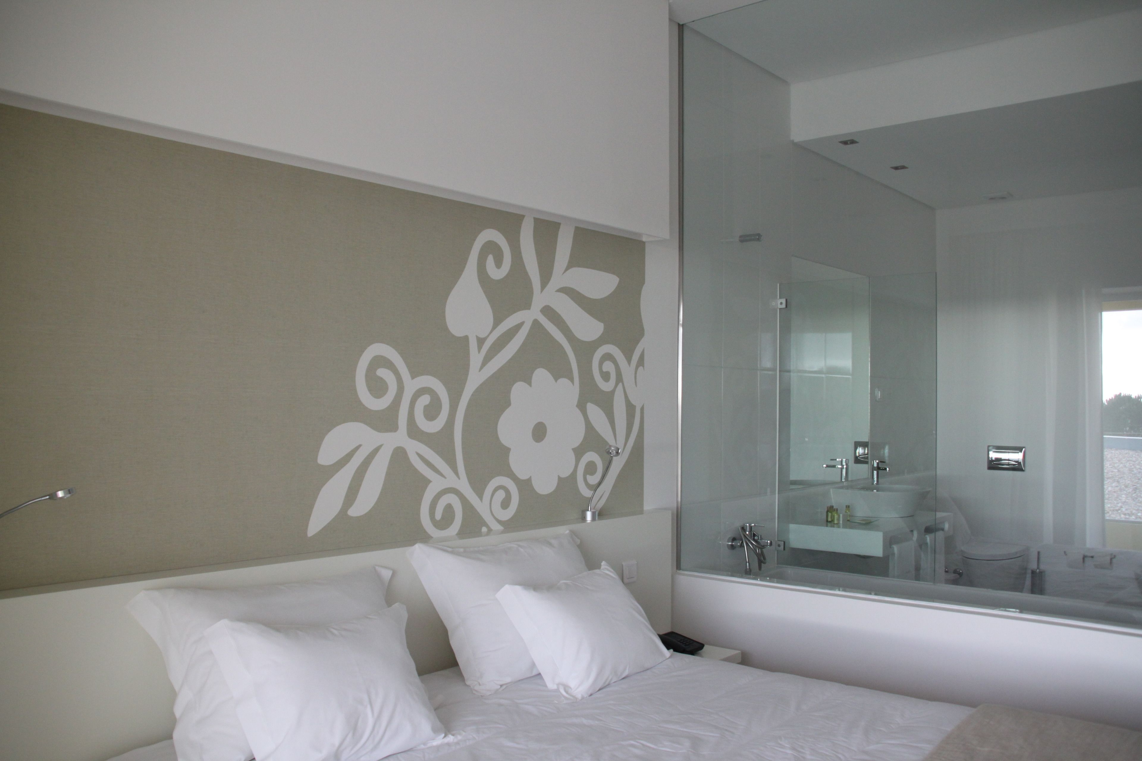 Un dormitorio moderno con una cama grande, una mesita de noche, un espejo y un baño visible a través de la puerta de cristal.