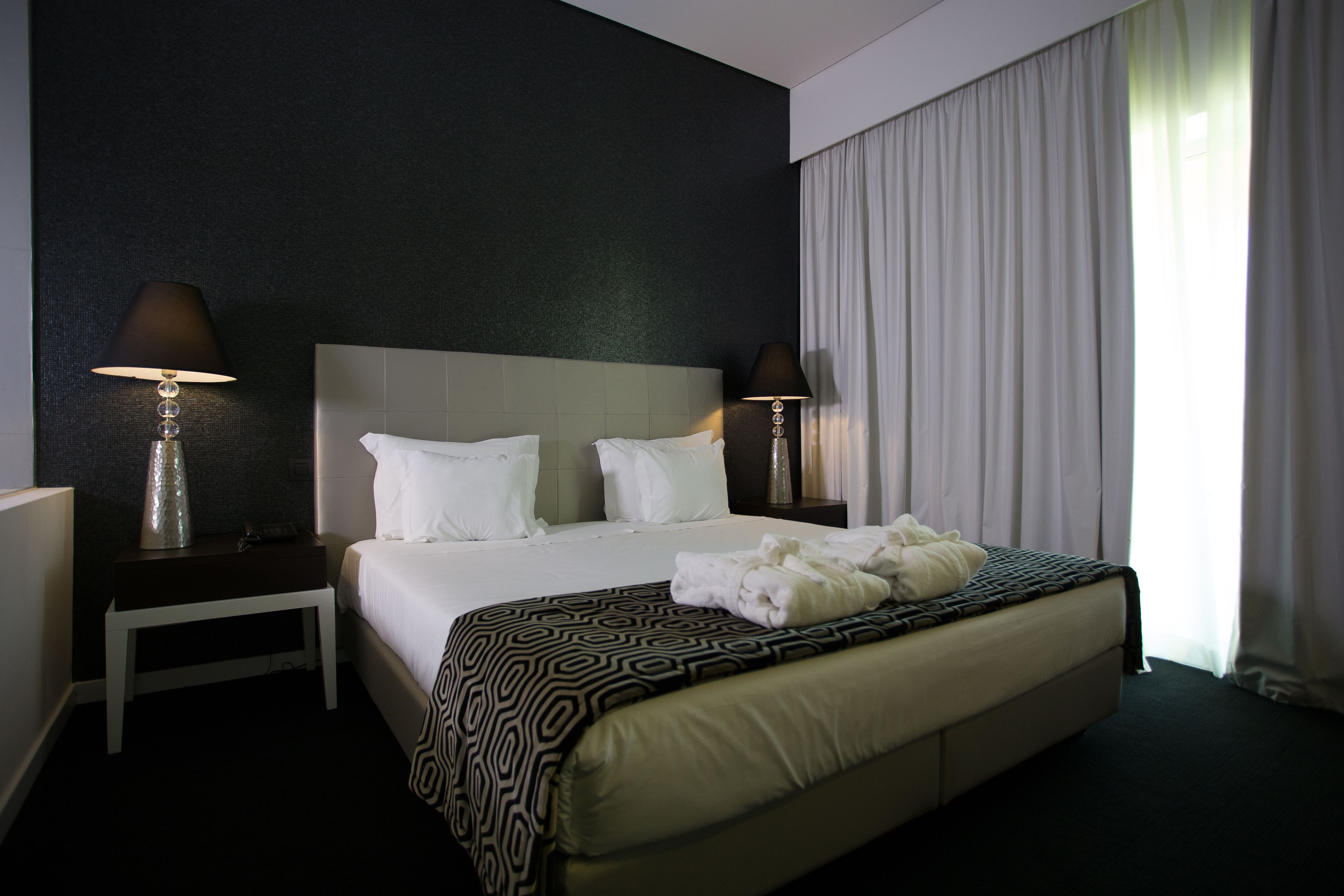Een hotelkamer met een groot bed, twee nachtkastjes met lampen, een hoofdbord en een raam met gordijnen.