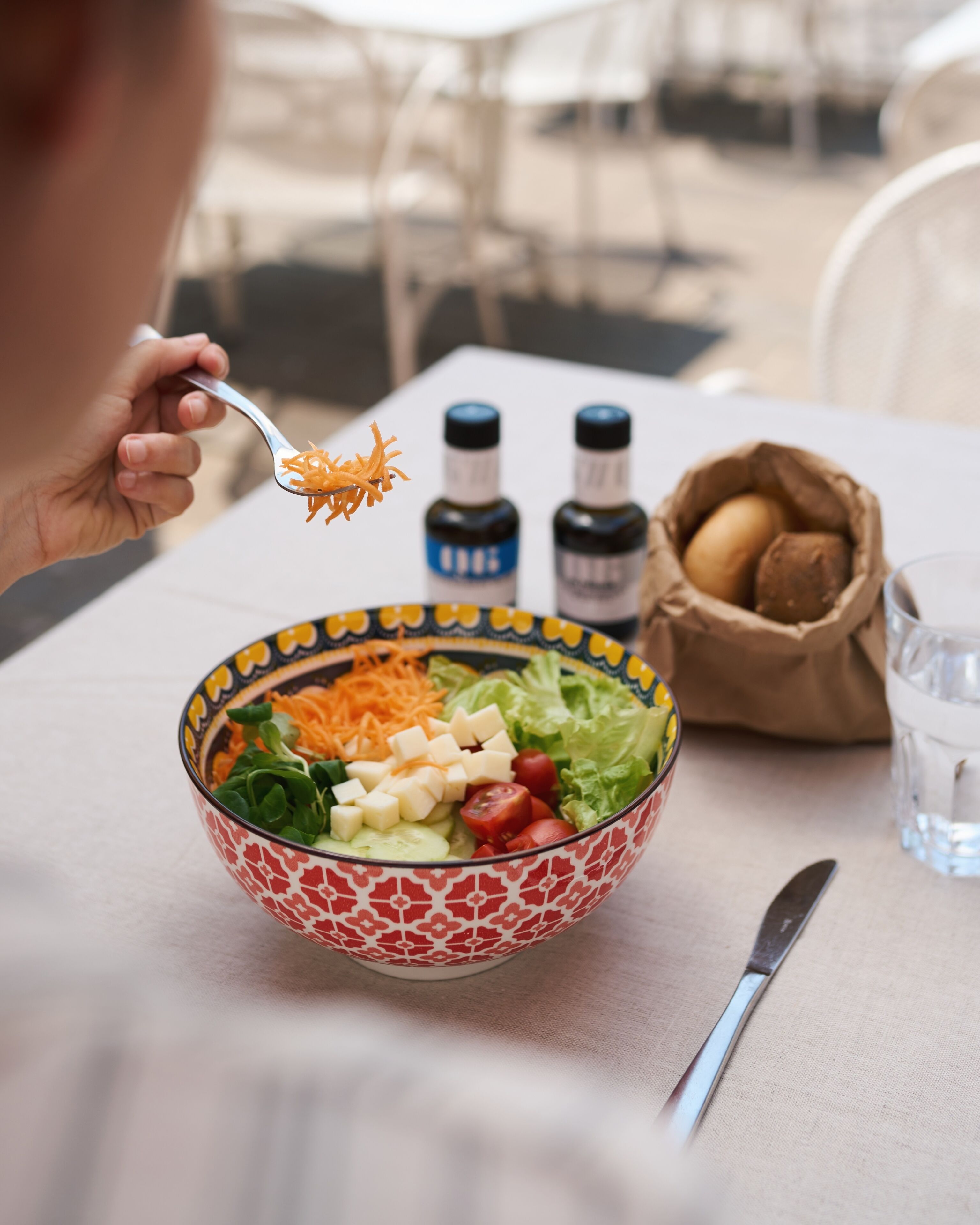 A bowl of salad with various vegetables and cheese, a knife, a glass of water, and two bottles of seasoning on a table.