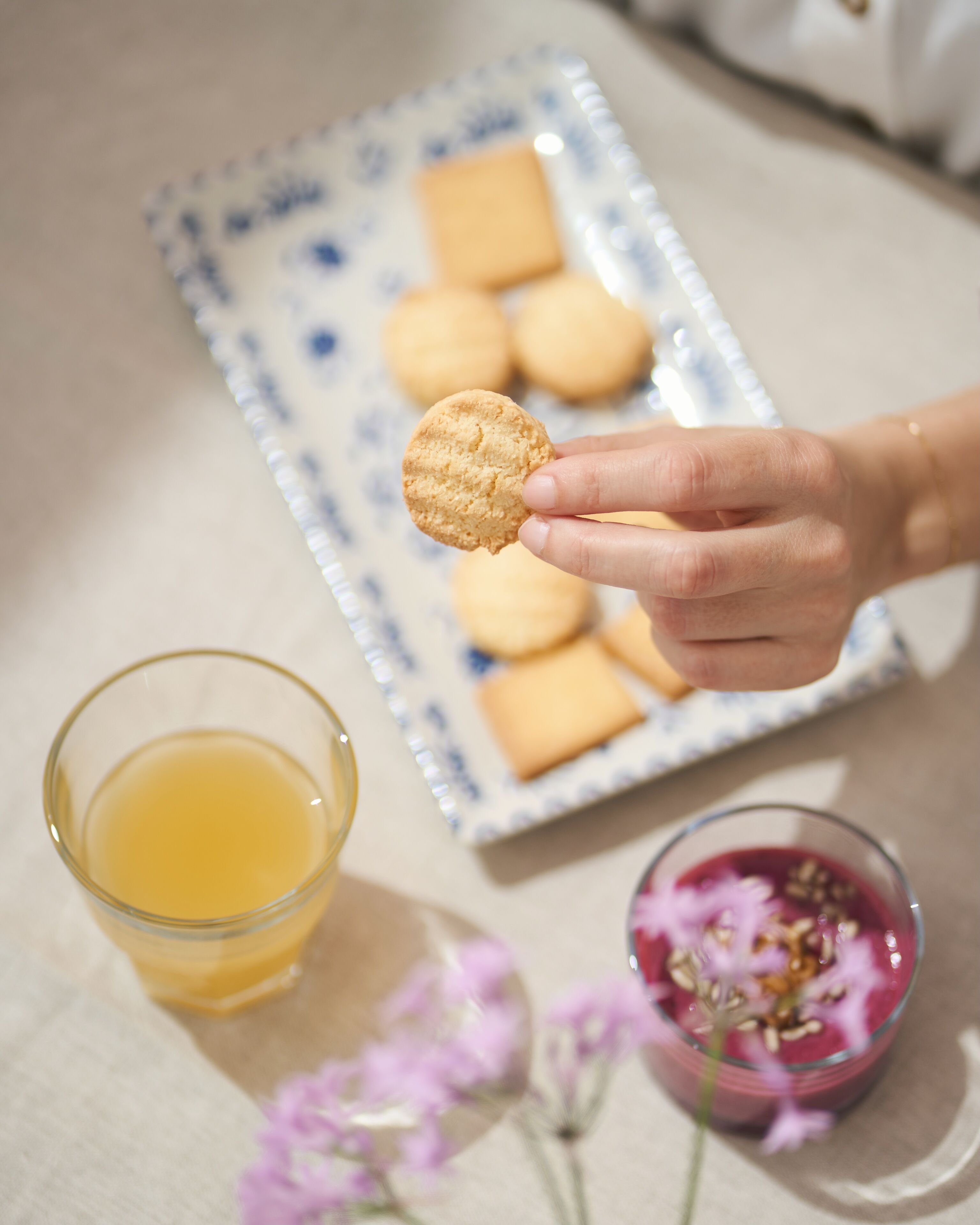 A hand holding a cookie, a glass of orange juice, and a plate with cookies in the background.