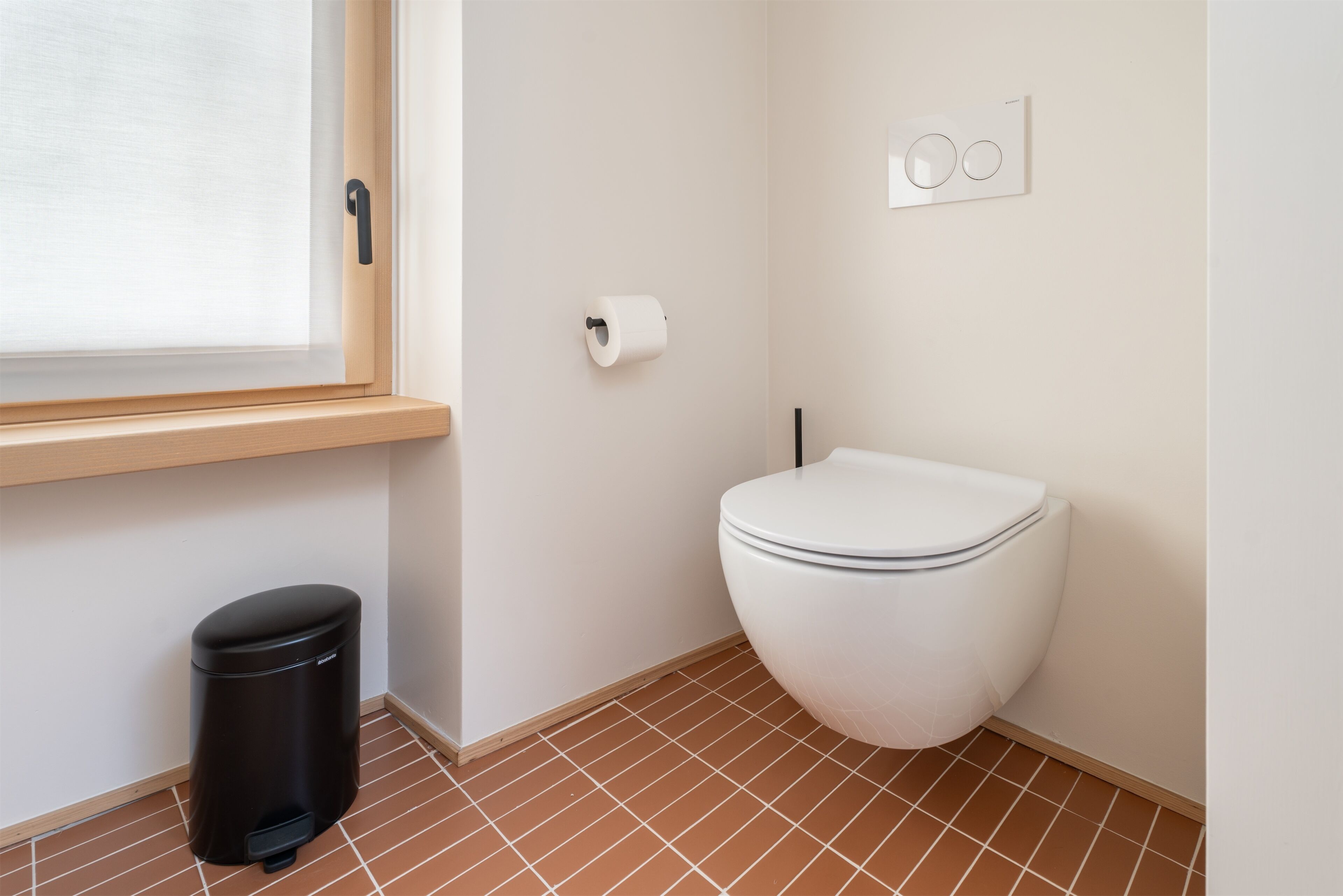 Un baño pequeño y limpio con inodoro, un cubo de basura, una ventana y un accionador de descarga en la pared.