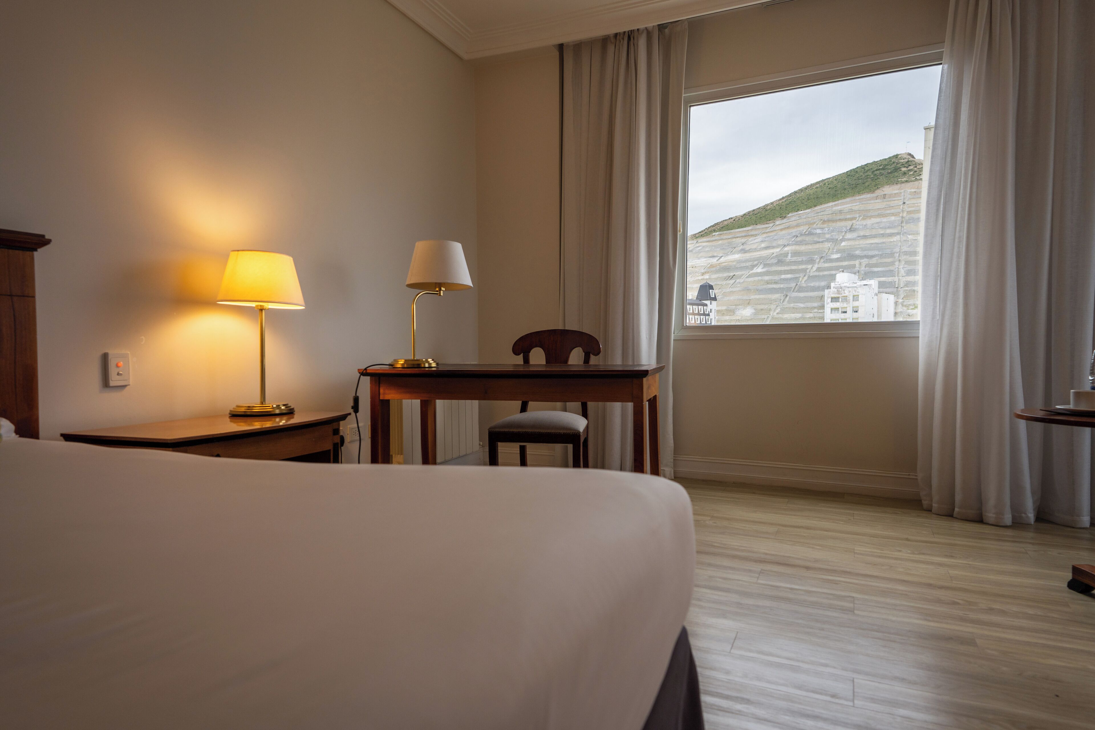 A hotel room with a bed, a desk, a chair, a lamp, and a window with a view of a building and hill.