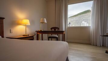 A hotel room with a bed, a desk, a chair, a lamp, and a window with a view of a building and hill.