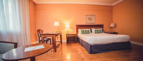 Habitación de hotel con piso de madera, cama con cubrecolcha azul, escritorio con silla y dos lámparas de noche.