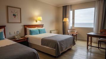A hotel room with two beds, a desk, and a view of the sea.