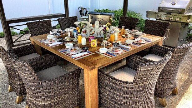 Une table de salle à manger en bois, entourée de chaises en osier, avec un barbecue et une clôture blanche à proximité.