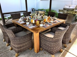 Una mesa de madera para comer con sillas de mimbre, rodeada por una parrilla y una valla blanca.