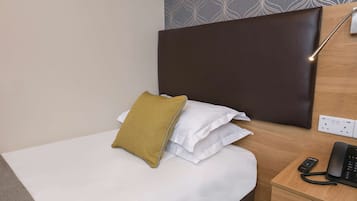 A neatly made bed with white and mustard pillows, a wooden nightstand with a black telephone, and a wall socket.