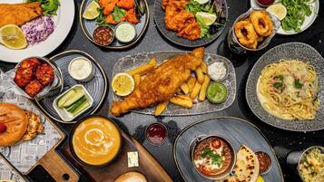 Variedad de platos, incluyendo pescado con papas fritas, hamburguesas y pasta, servidos en una mesa con periódicos como posavasos.