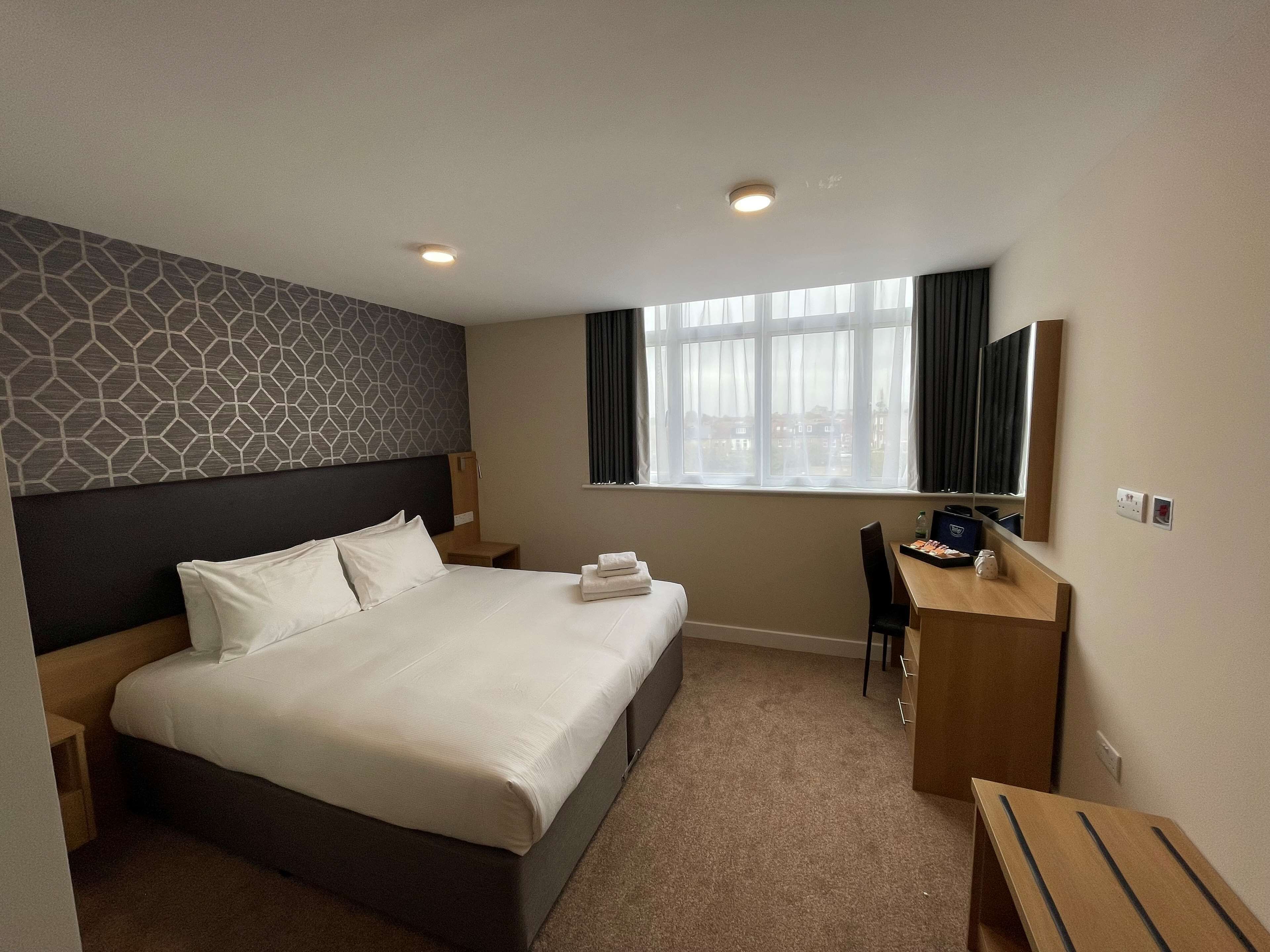 A hotel room with a large bed, a desk, a chair, a small table, a window with curtains, and a patterned wall.