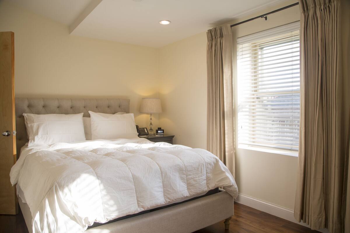 A bedroom with a large bed, a bedside table with a lamp, and a window with curtains.