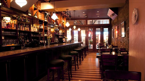 Un bar con una barra larga, varios taburetes y piso a rayas.
