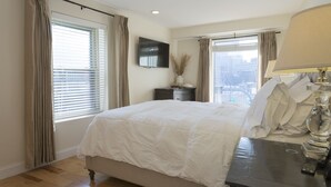 Un dormitorio con una cama grande, una mesita de noche con lámpara, un televisor montado en la pared y una ventana con cortinas.