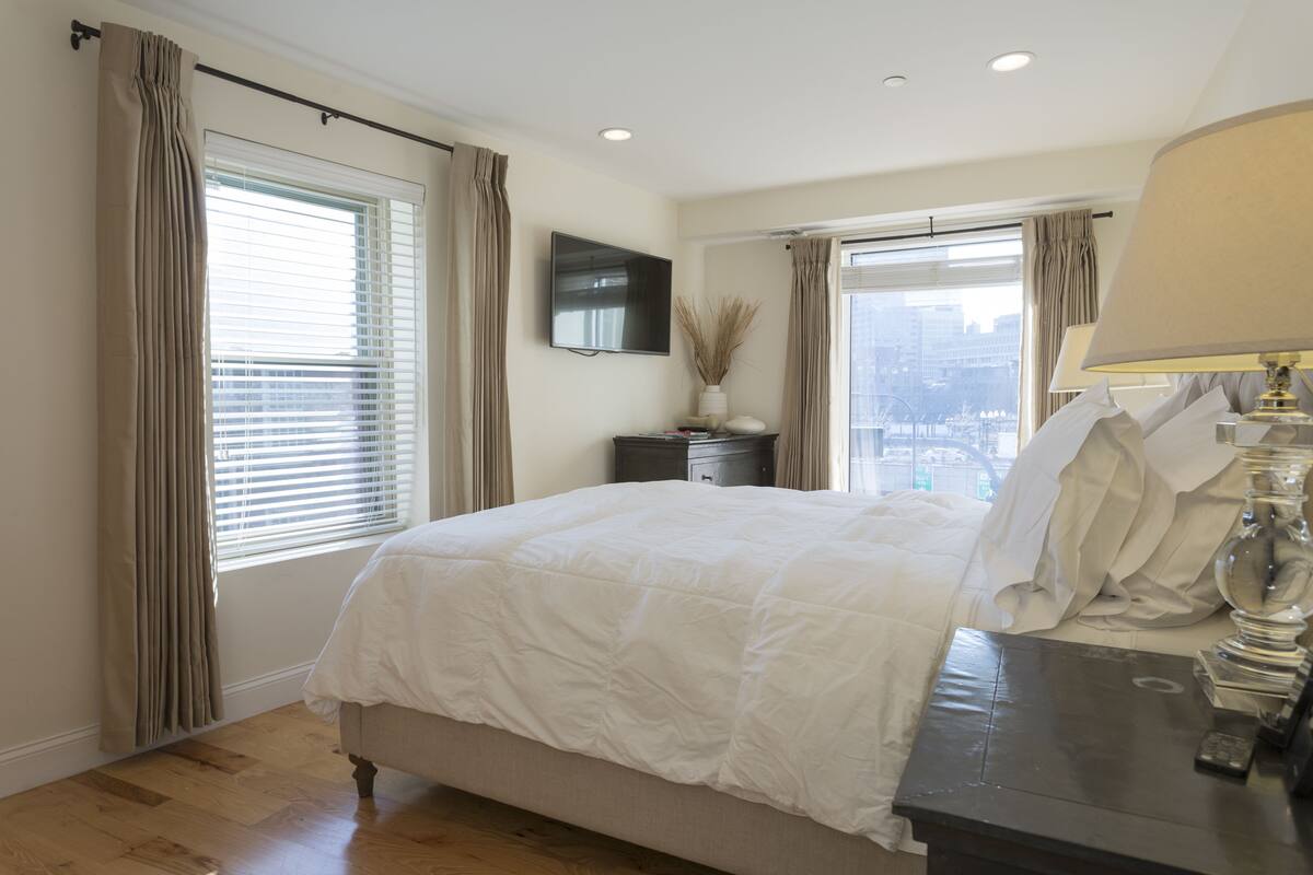 A bedroom with a large bed, a bedside table with a lamp, a TV mounted on the wall, and a window with curtains.