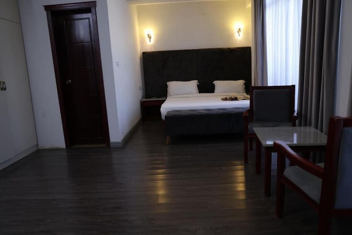 Habitació d'hotel amb sòl de fusta fosca, llit amb roba blanca, una nitòria, escriptori amb cadira i una porta.