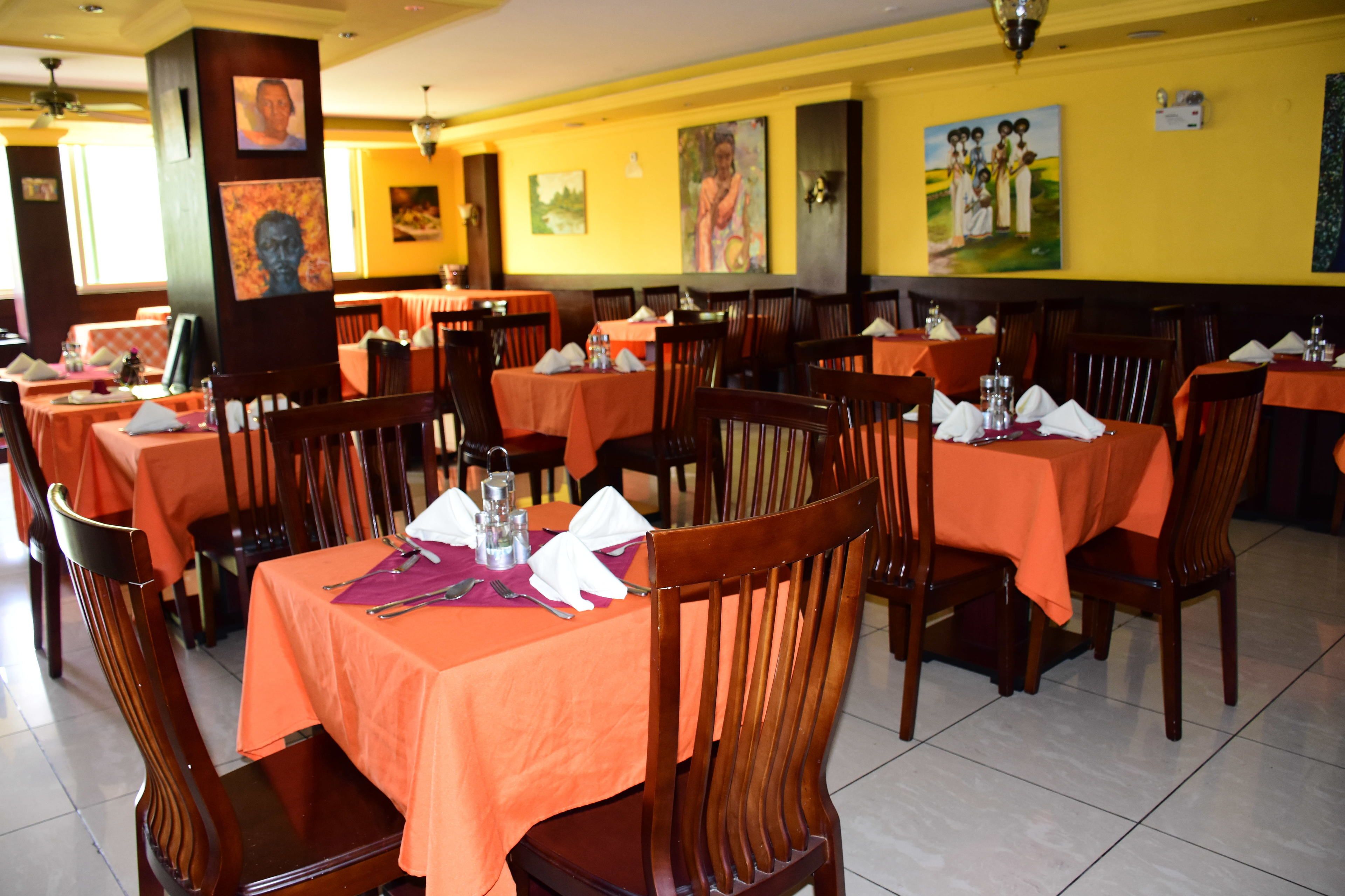 A restaurant with tables covered in orange tablecloths, wooden chairs, and paintings on the walls.