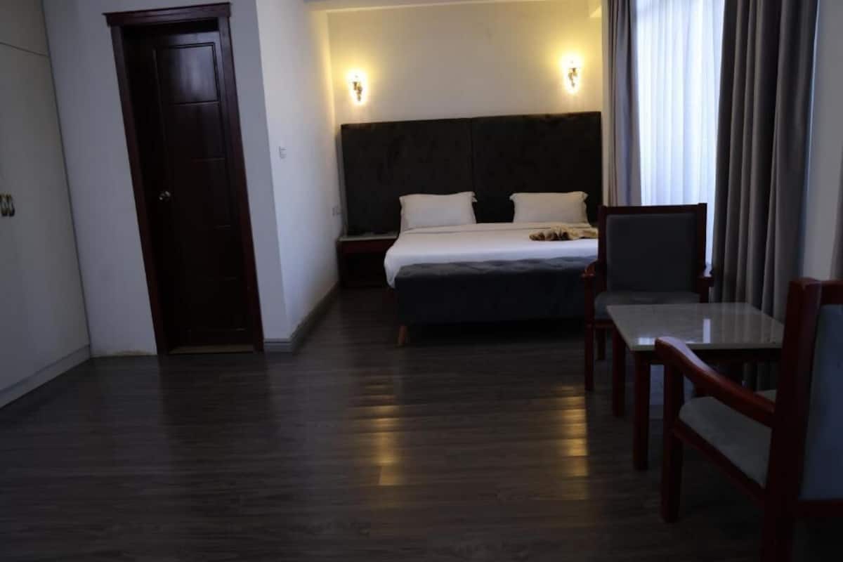 A hotel room with a dark wood floor, a bed with white linens, a wooden nightstand, a desk with a chair, and a door.