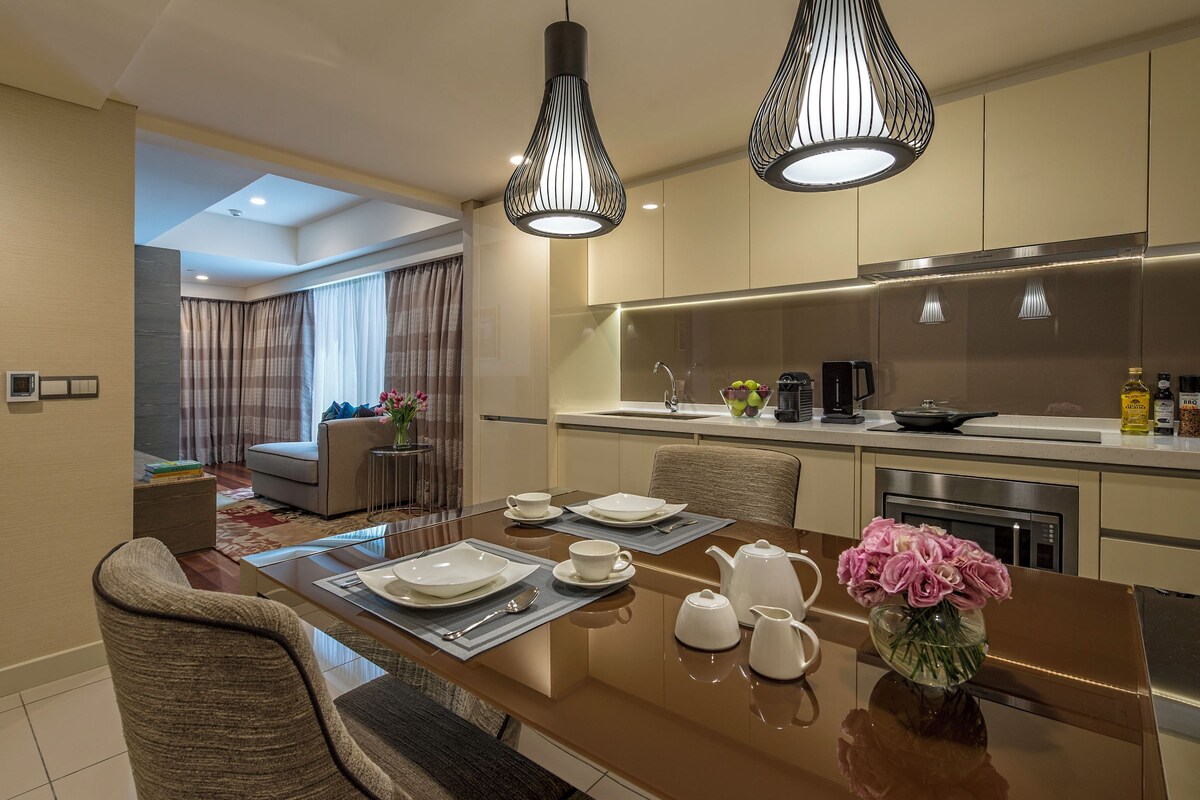 A modern dining area with a long table, chairs, and a vase of flowers. There is a kitchen area with a sink, stove, and cabinets.