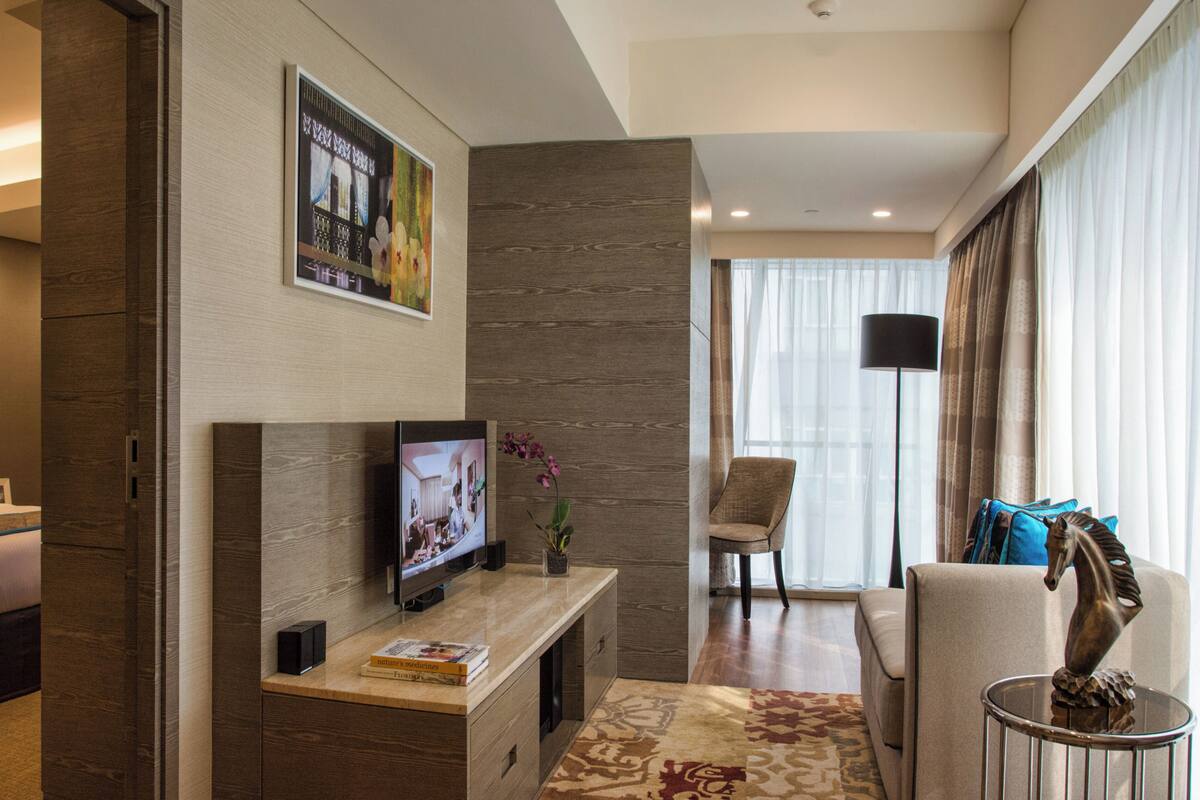 A hotel room with a bed, a TV on a wooden console, a chair, a sofa, and a large window with curtains.