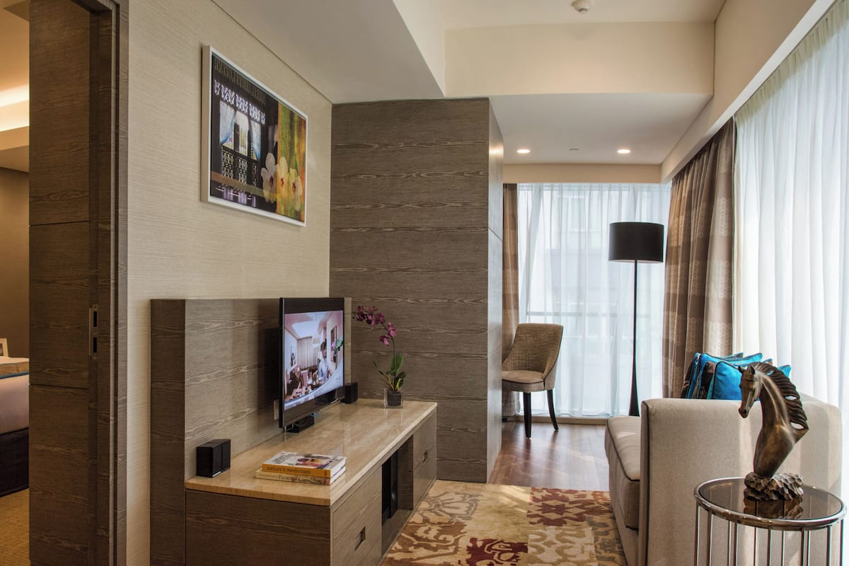 Una habitación de hotel con una cama, un televisor sobre un mueble de madera, una silla, un sofá y un ventanal con cortinas.