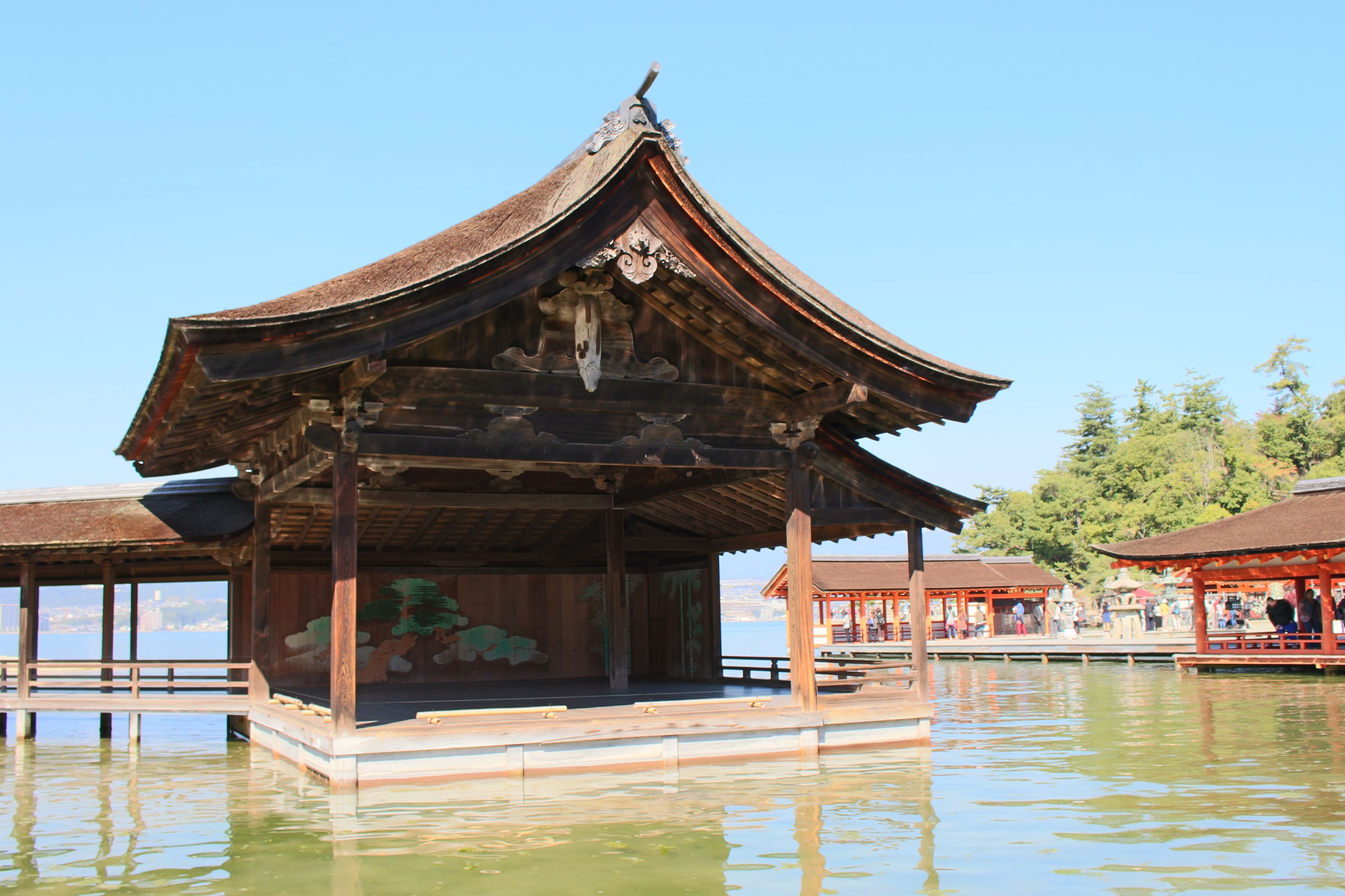 Ein traditioneller, hölzerner Pavillon auf Stelzen über dem Wasser, mit blauem Himmel im Hintergrund.