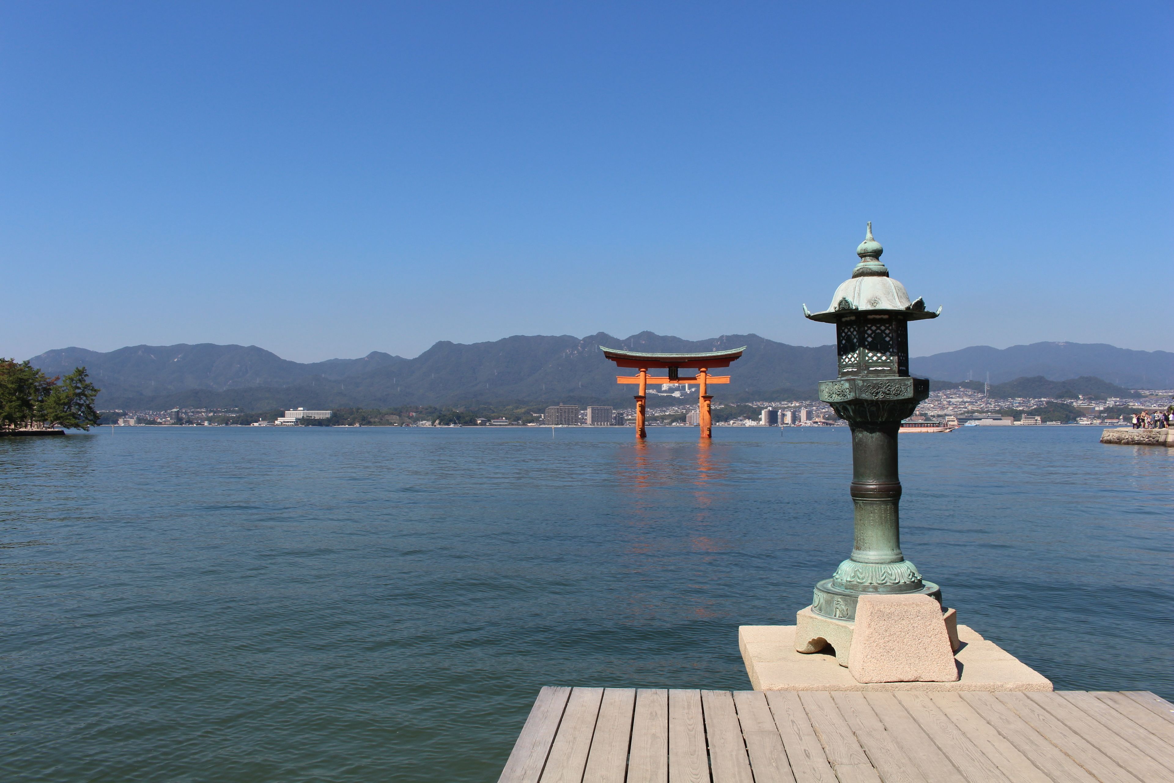 Un muelle de madera se adentra en un cuerpo de agua tranquilo, con un torii japonés tradicional al fondo y montañas a lo lejos.