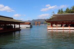 A traditional Japanese building with a thatched roof, a water body in the foreground, and mountains in the background. - Miyajima Seaside Hotel (Hatsukaichi)