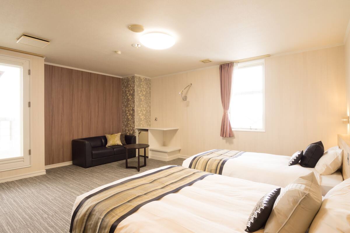 Habitación de hotel con dos camas, un sofá, una mesita, una lámpara y una ventana con cortinas.