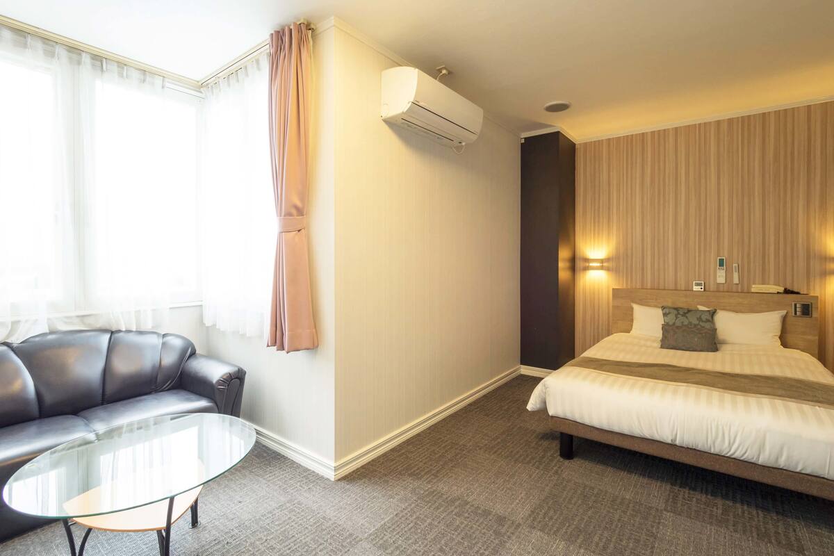 Una habitación de hotel con una cama, un sofá de cuero, una mesita de centro de vidrio y un aire acondicionado instalado en la pared.